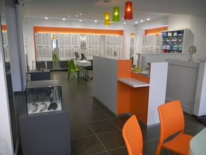 Intérieur magasin d'optique agencé avec présentoirs à lunette au mur du fond, banque d'accueil en blanc et orange au centre, vitrine à gauche et chaises orange tout devant