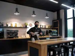 intérieur showroom Cafés Richard. Un homme habiller de noir prépare le café sur une table en bois de chêne avec incrustation solid surface noir. Derrière un meuble bas noir avec dessus bois clair et machines à cafés. Mur du fond blanc avec étagères noires.