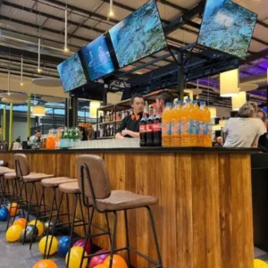 Bar central dans un centre de loisirs avec chaises hautes devant le comptoir, des serveur à l'arrière, et un arrière-bar avec un rayonnage métallique et 4 grands écrans en hauteur.