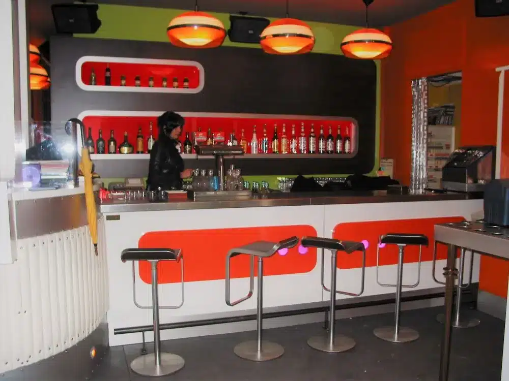 Bar style années 70 comptoir blanc avec motifs oranges dessus en inox et arrière bar avec niche retro-éclairées orange et suspentes forme ovoÏde orange translucide, une personne derrière de comptoir habillée de noir.