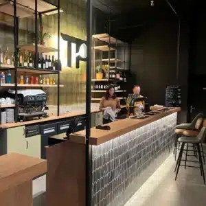 Bar moderne avec comptoir avec dessus en bois clair et façade avec carrelage vert, 1 homme et 1 femme derrière le comptoir, arrière bar avec frigo froid et rayonnage métallique, enseigne avec initiale TP retro-éclairée au centre