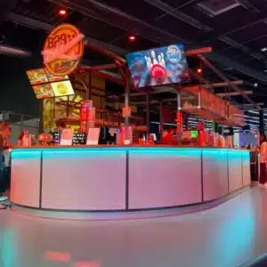 Bar cintré façade éclairée par led bleu avec grille en métal déployé, dessus bar en bois, arrière bar en structure métallique avec écrans en hauteur.