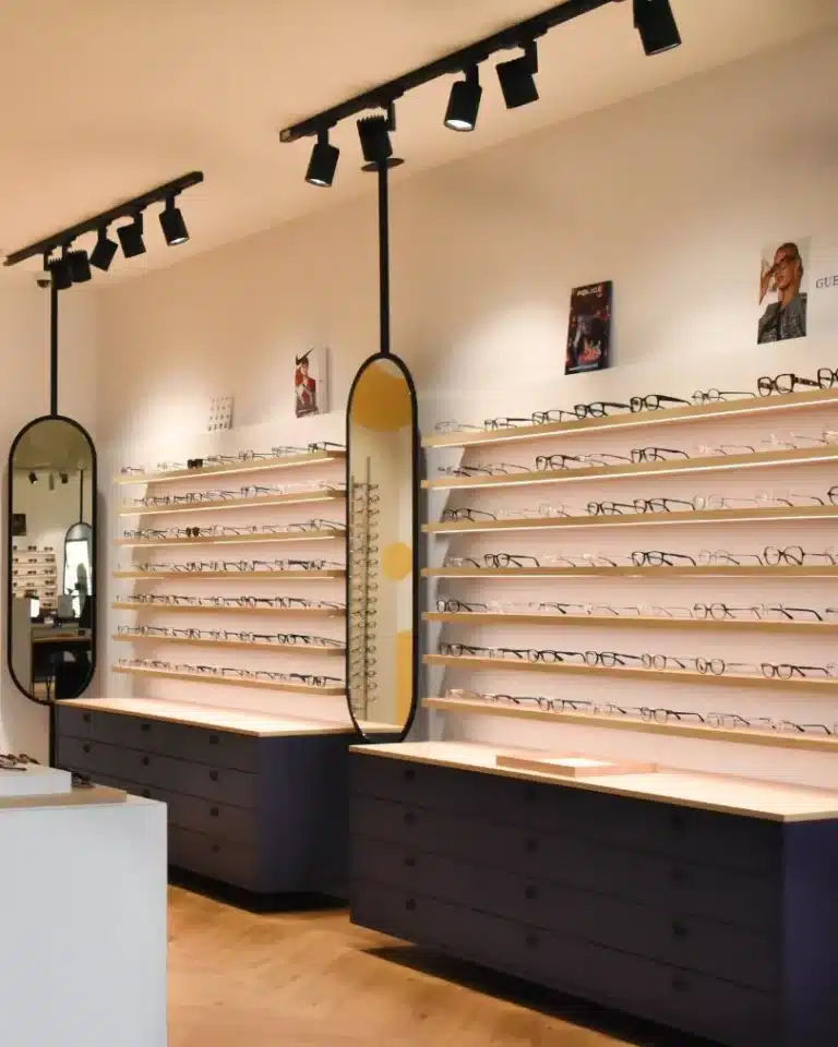 Agencement intérieur magasin d'optique avec murs blancs, sol plancher bois clair, 2 colonnes de 7 étagères suspendues au mur avec lunettes rétro-éclairées, 2 meubles laqués bleu foncé suspendus au murs, 2 miroirs arrondis