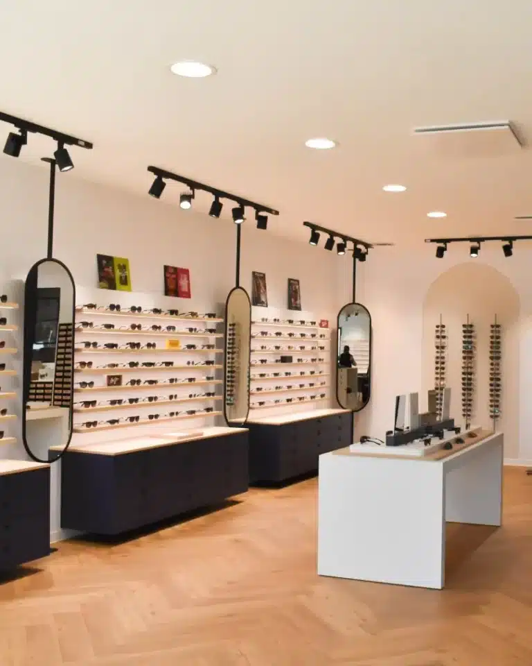 Intérieur optique avec table de présentation et présentoirs à lunettes suspendus aux murs avec 3 miroirs.