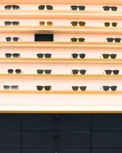 Etageres en bois clair avec LED incrusté suspendues au mur blanc avec plusieurs modèles de lunettes et meuble bas avec tiroirs laqués bleu et poignée noire