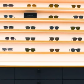 Etageres en bois clair avec LED incrusté suspendues au mur blanc avec plusieurs modèles de lunettes et meuble bas avec tiroirs laqués bleu et poignée noire