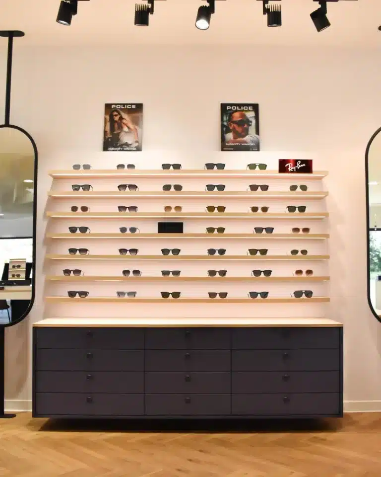 Sept étagères en bois clair suspendues au mur blanc avec 6 modèles de lunettes chacune. Meuble bas laqué bleu foncé suspendu en partie basse. 2 miroirs ovales sur les côtés