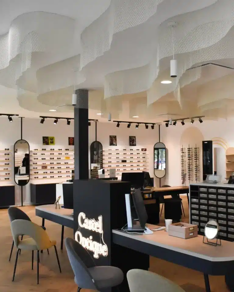 Intérieur magasin d'optique avec table de vente et 4 fauteuils devant, présentoirs à lunettes suspendus à l'arrière plan le long des murs, voiles décoratifs au plafond.